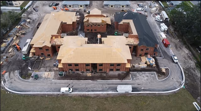 Aerial view of a large building under construction with multiple sections and a surrounding paved road. Construction materials and vehicles are visible around the site.