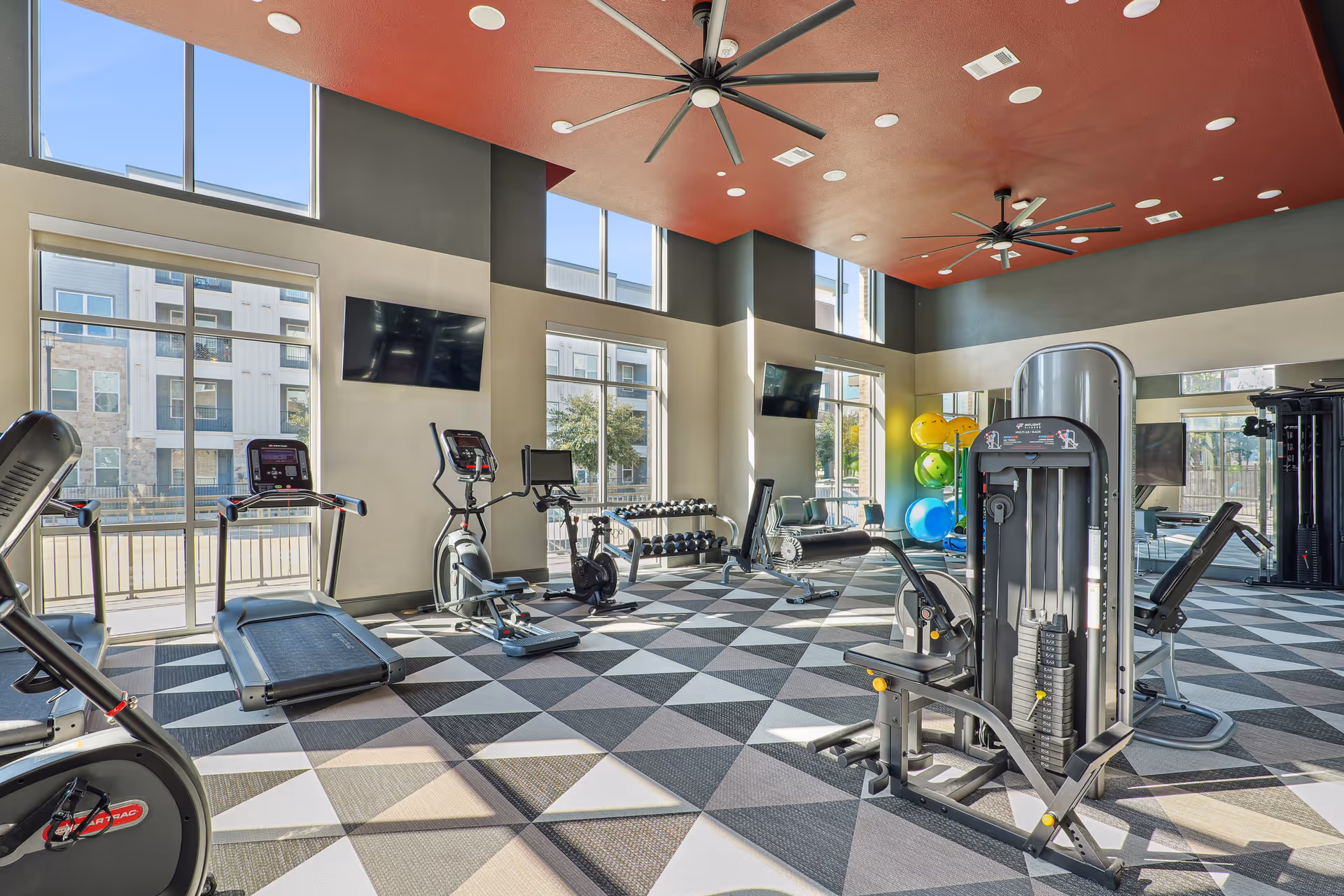 Bright and spacious fitness center with large windows, exercise machines including treadmills, stationary bikes, weight machines, and a rack of colorful exercise balls. The room has a patterned floor, red ceiling with large ceiling fans, and multiple wall-mounted TVs.
