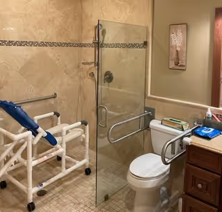 Accessible bathroom with a glass-enclosed shower, grab bars, a rolling shower chair, and a toilet beside a vanity.