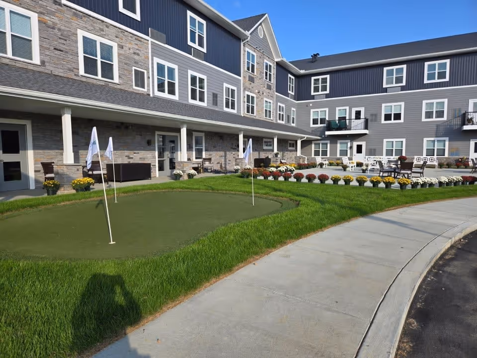Outdoor view of a senior living facility named The Residence at Village Greens featuring a small putting green with flags, a curved sidewalk, flower beds with colorful flowers, and a multi-story building with balconies and windows in the background.