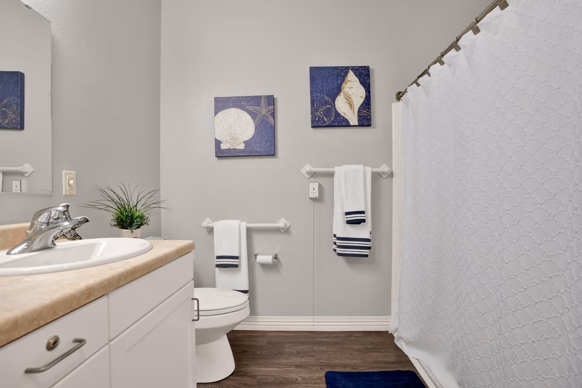 A clean bathroom with a white sink and countertop on the left, a toilet in the center with two white towels hanging on towel racks above it, and a white shower curtain on the right. The walls are light gray with two blue and white seashell-themed pictures hanging above the towels. There is a small green plant on the countertop and a dark blue bath mat on the floor.