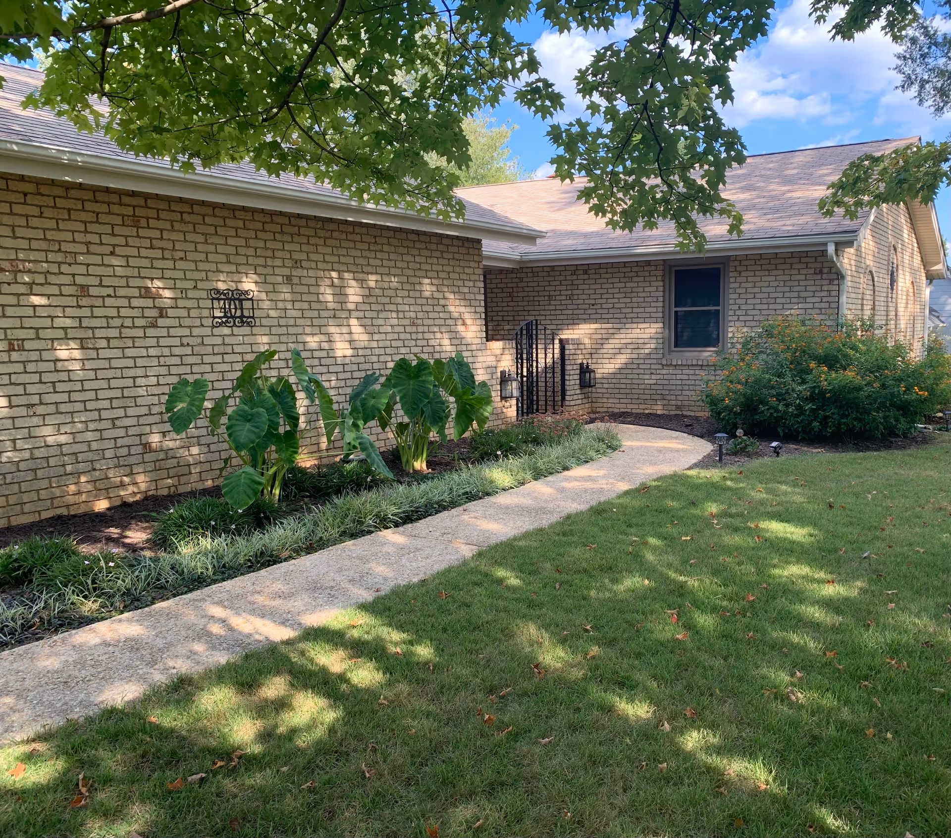 A brick building with a curved concrete pathway leading to a black metal gate. The building is surrounded by green grass, bushes, and plants, with tree branches and leaves partially shading the scene under a blue sky with some clouds.