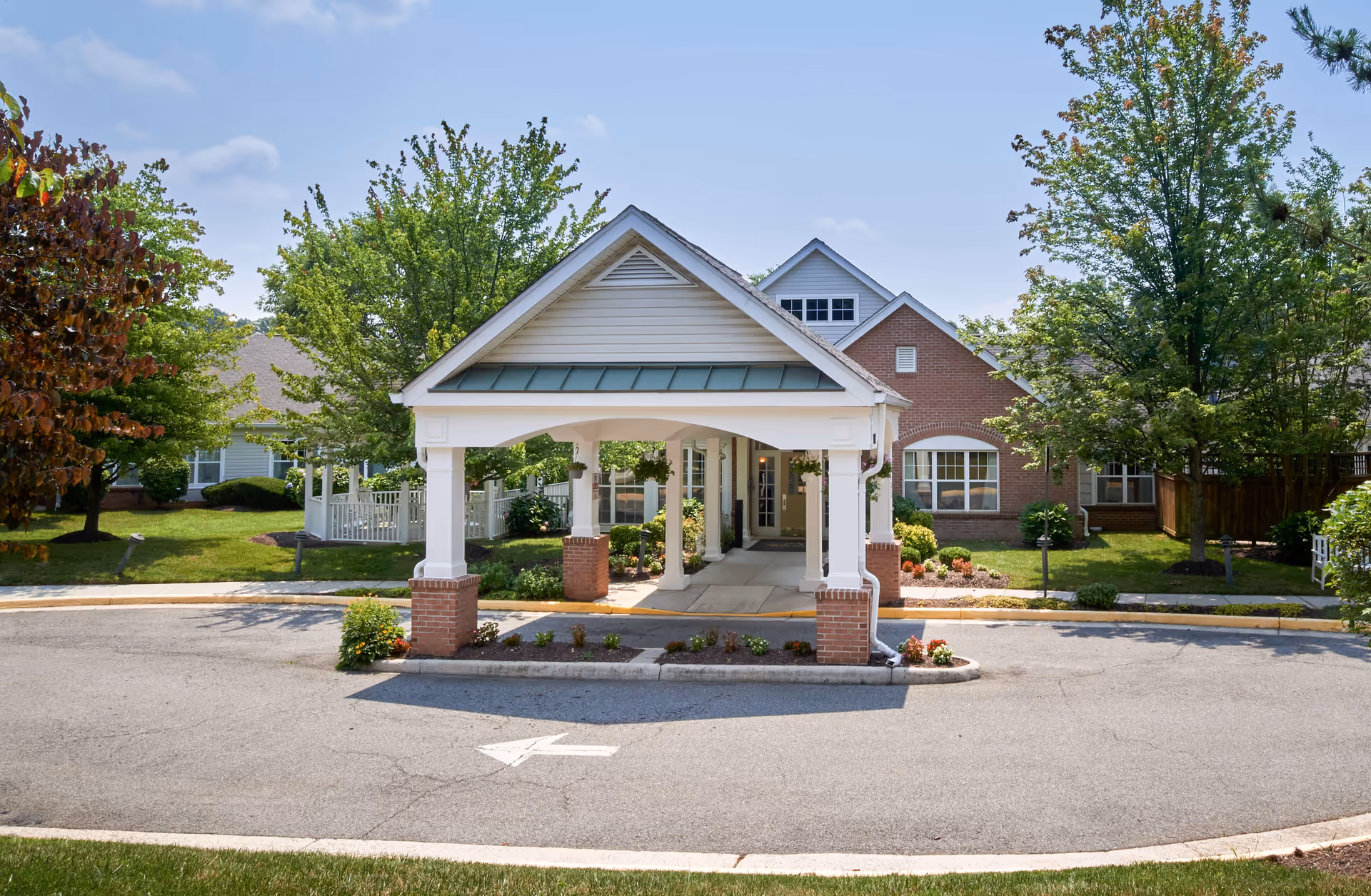 Entrance to a senior living facility with a covered drop-off area supported by white columns with brick bases. The building has a mix of brick and siding exterior, surrounded by green trees and landscaping under a clear blue sky.