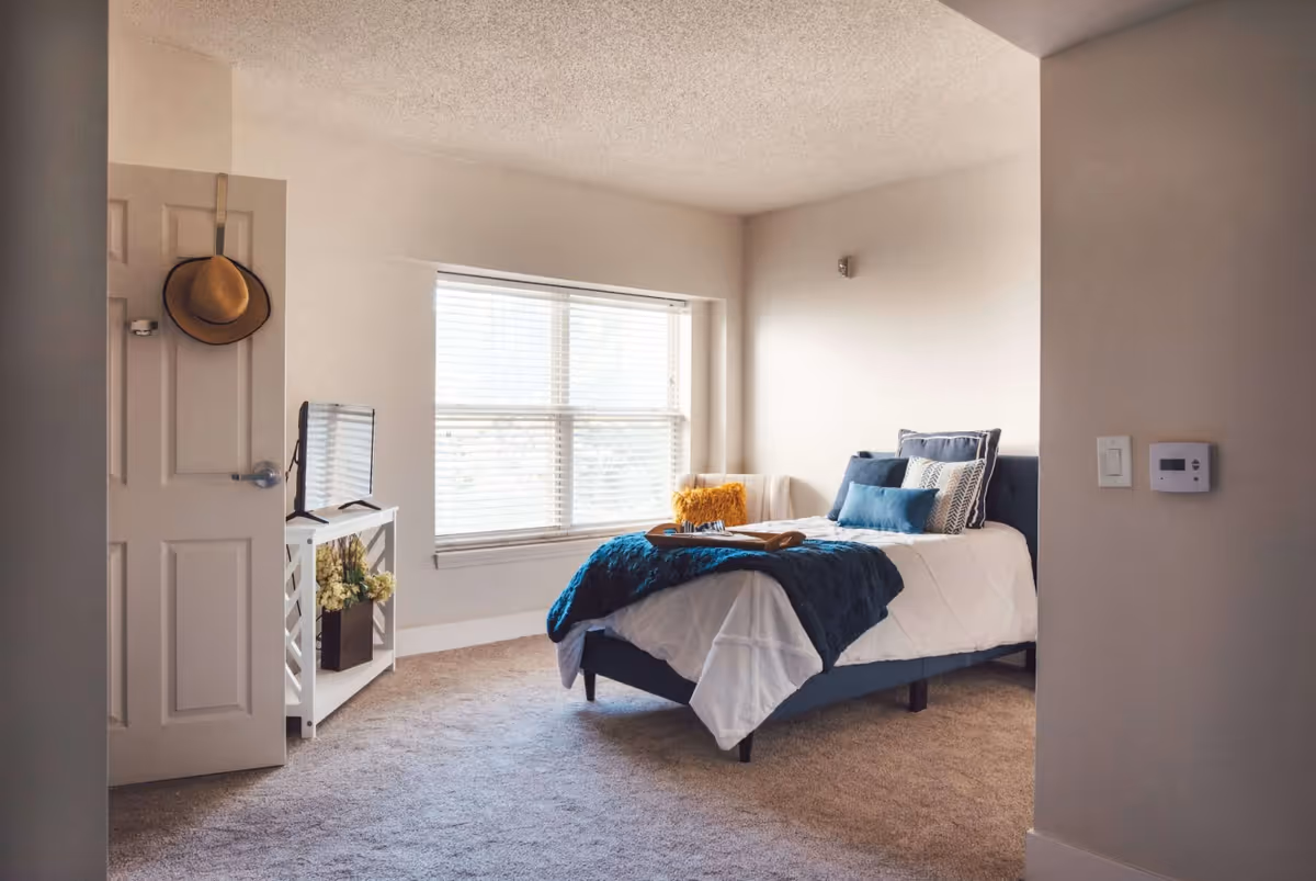 A cozy bedroom with a single bed dressed in white and blue bedding, several decorative pillows, and a tray on the bed. There is a large window with blinds letting in natural light, a small white TV stand with a flat-screen TV and a vase of flowers, and a door with a hat hanging on it. The room has beige walls and carpeted floor.