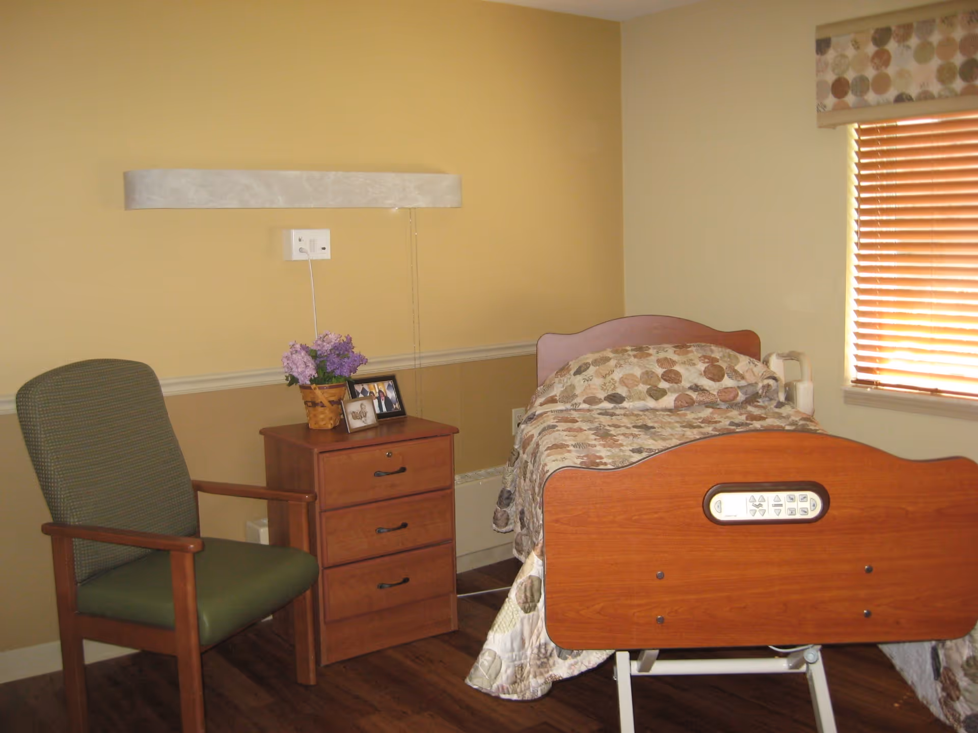 A furnished senior bedroom with a medical-style wooden bed, nightstand holding flowers and framed photos, and a chair beside a window with blinds.
