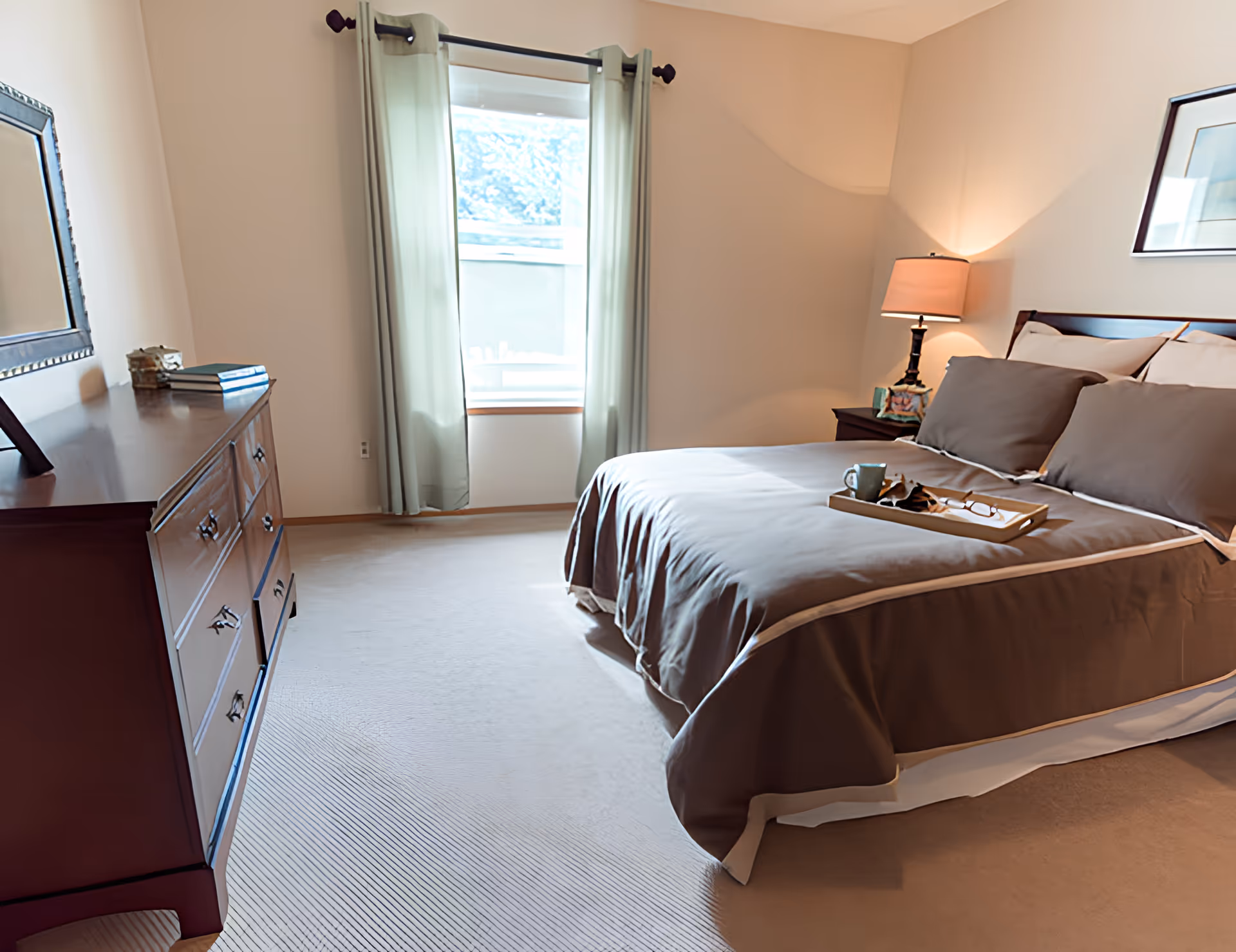 A neatly made bed with brown bedding and pillows in a bedroom. A wooden dresser with books and decorative items is on the left side. A window with light green curtains allows natural light into the room. A bedside table with a lamp and a framed picture is on the right side.