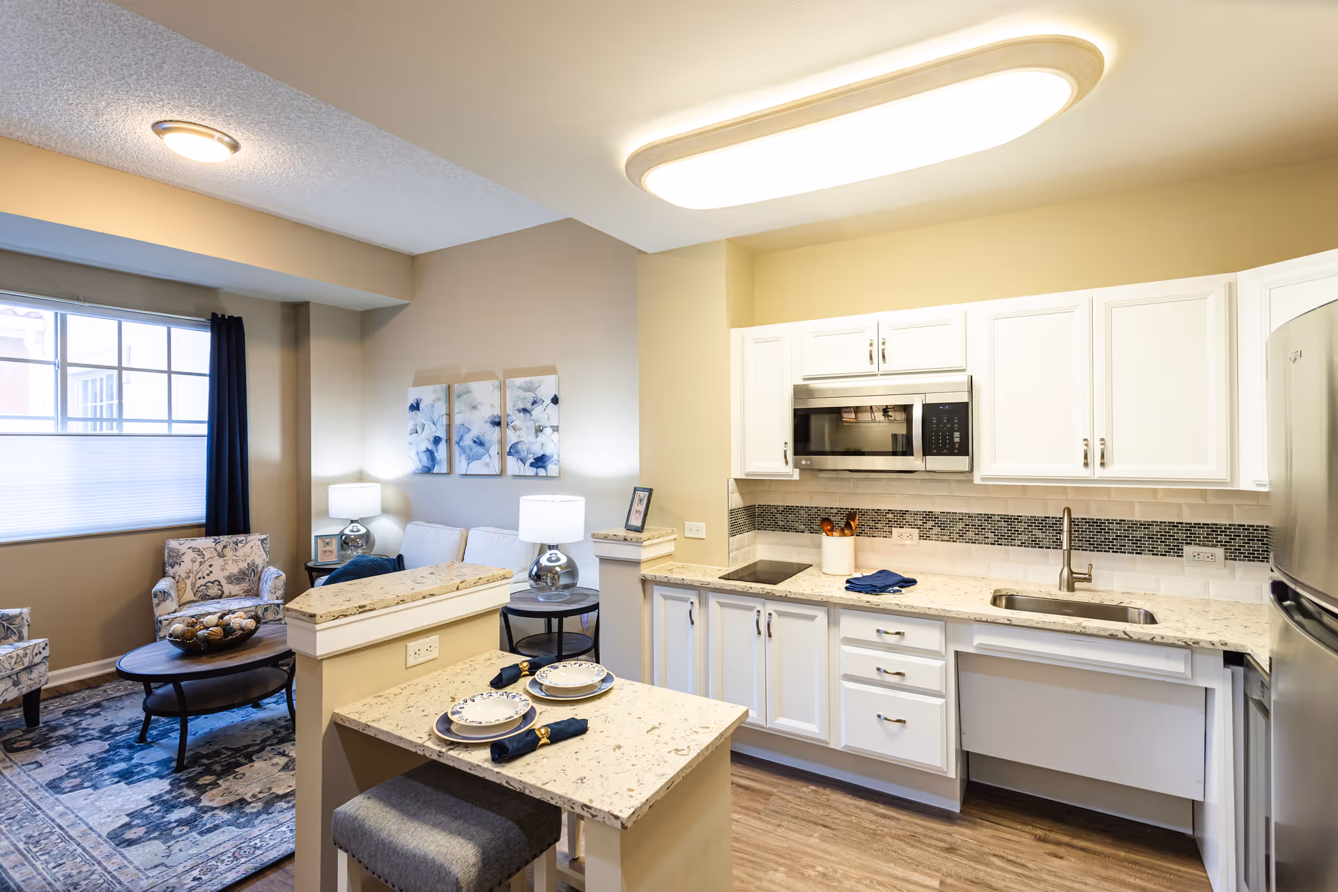 A bright and modern senior living apartment interior featuring a kitchen and living room area. The kitchen has white cabinets, a stainless steel microwave and refrigerator, a sink, and a small island with two place settings. The living room includes two patterned armchairs, a coffee table, a sofa, two table lamps, and three floral wall art pieces. A large window with dark curtains allows natural light into the space.