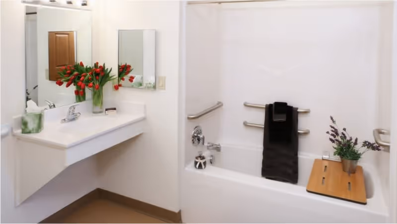 A clean and accessible bathroom featuring a white sink with a silver faucet, a large mirror above the sink, and a vase of red tulips on the counter. The bathtub has multiple grab bars for safety, a black towel hanging on one of the bars, and a wooden bath tray with a small potted plant on it.