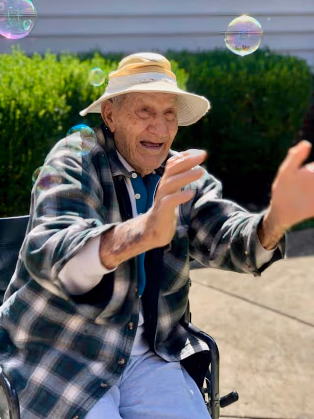 An elderly man wearing a wide-brimmed hat and a plaid shirt is sitting in a wheelchair outdoors, joyfully reaching out to catch bubbles floating around him with green bushes and a building wall in the background.