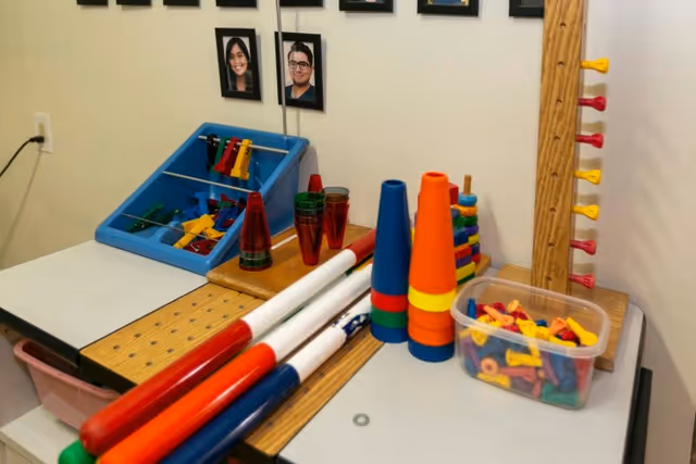 A table with various colorful therapy and activity tools including stacking cones, plastic cups, rods, and pegs in a room with framed photos on the wall.