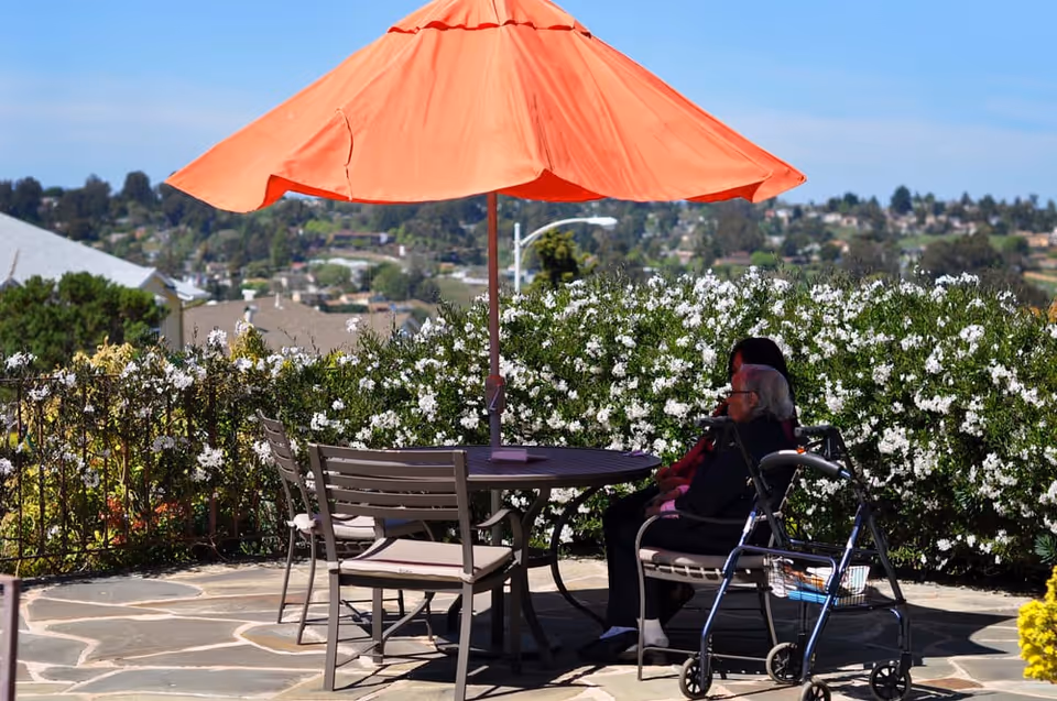 Two elderly individuals sitting at a round outdoor table under a large orange umbrella on a stone patio. One person is seated in a wheelchair, and there is a walker nearby. The background shows green bushes with white flowers and a distant view of houses and trees under a clear blue sky.