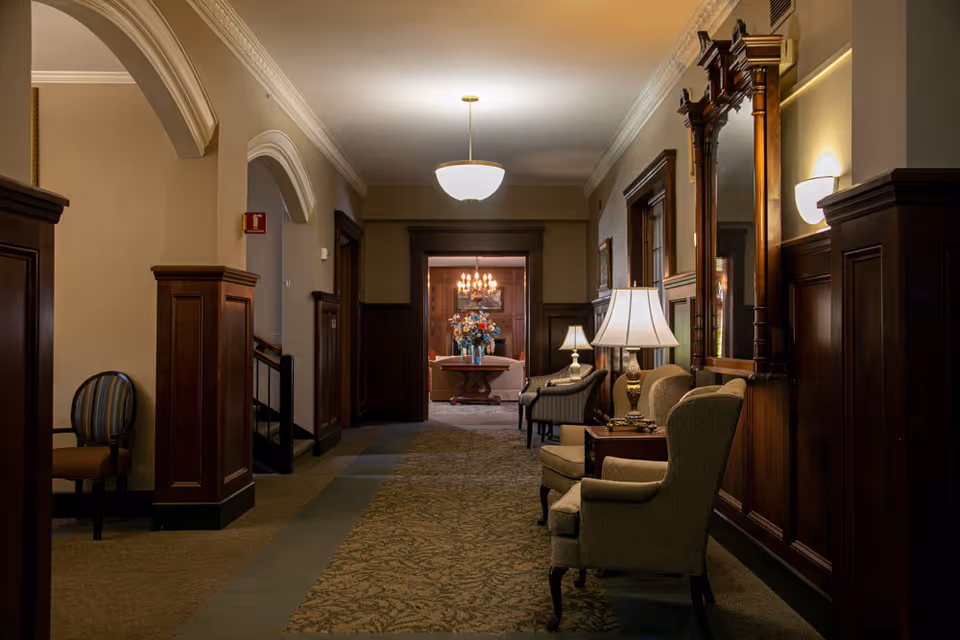 A warmly lit hallway in a senior living facility with wooden paneling on the walls, upholstered chairs and side tables with lamps along the right side, and a carpeted floor. At the end of the hallway, there is an open doorway leading to a room with a chandelier, a floral arrangement on a table, and additional seating.