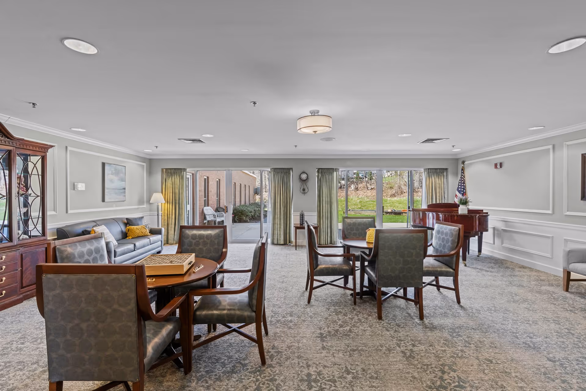 A spacious senior living common area with several round tables and chairs arranged for socializing or games. There is a gray sofa with yellow pillows, a wooden cabinet with glass doors, a floor lamp, and a grand piano in the corner. Large windows and glass doors provide a view of an outdoor patio and greenery. The room has light-colored walls with decorative molding and a carpeted floor.