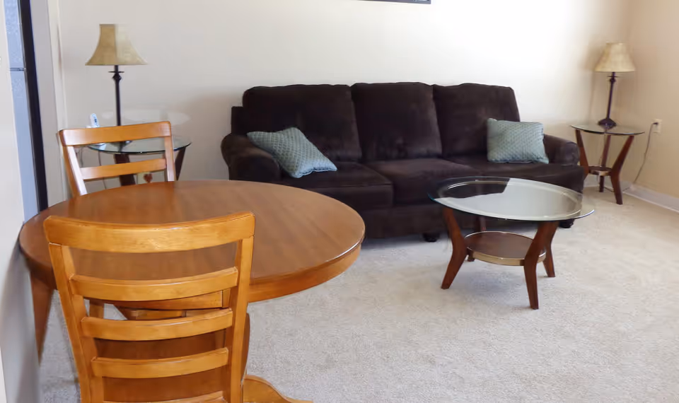 A living room area with a dark brown sofa adorned with two light blue pillows, a round wooden dining table with two wooden chairs, a glass-top coffee table with wooden legs, and two side tables each holding a lamp with a beige lampshade. The room has light-colored carpet and neutral walls.