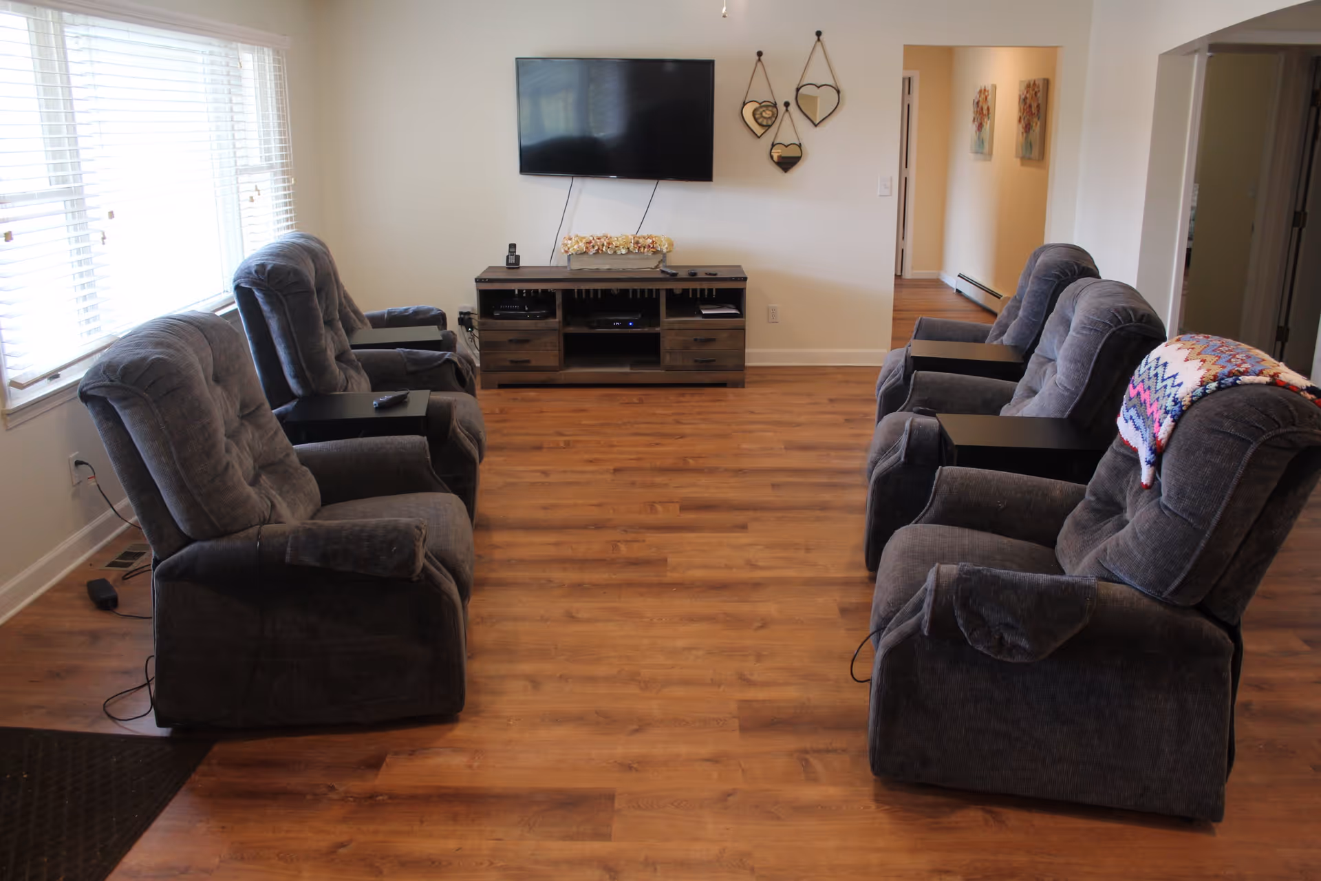 A living room with six gray recliner chairs arranged in two rows facing a wall-mounted flat screen TV. Below the TV is a wooden media console with decorative items and electronic devices. The room has wooden flooring, white walls, and a window with blinds on the left side. There are three heart-shaped mirrors hanging on the wall next to the TV and two floral paintings visible in a hallway to the right.