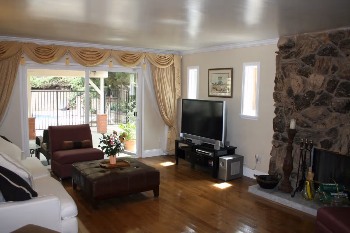 A cozy living room with wooden flooring, a white sofa, a maroon armchair, and a brown ottoman with a flower vase on top. There is a large flat-screen TV on a black TV stand, two small windows, and a stone fireplace with candles and fireplace tools. Sliding glass doors with gold curtains open to an outdoor area with a pool and greenery.