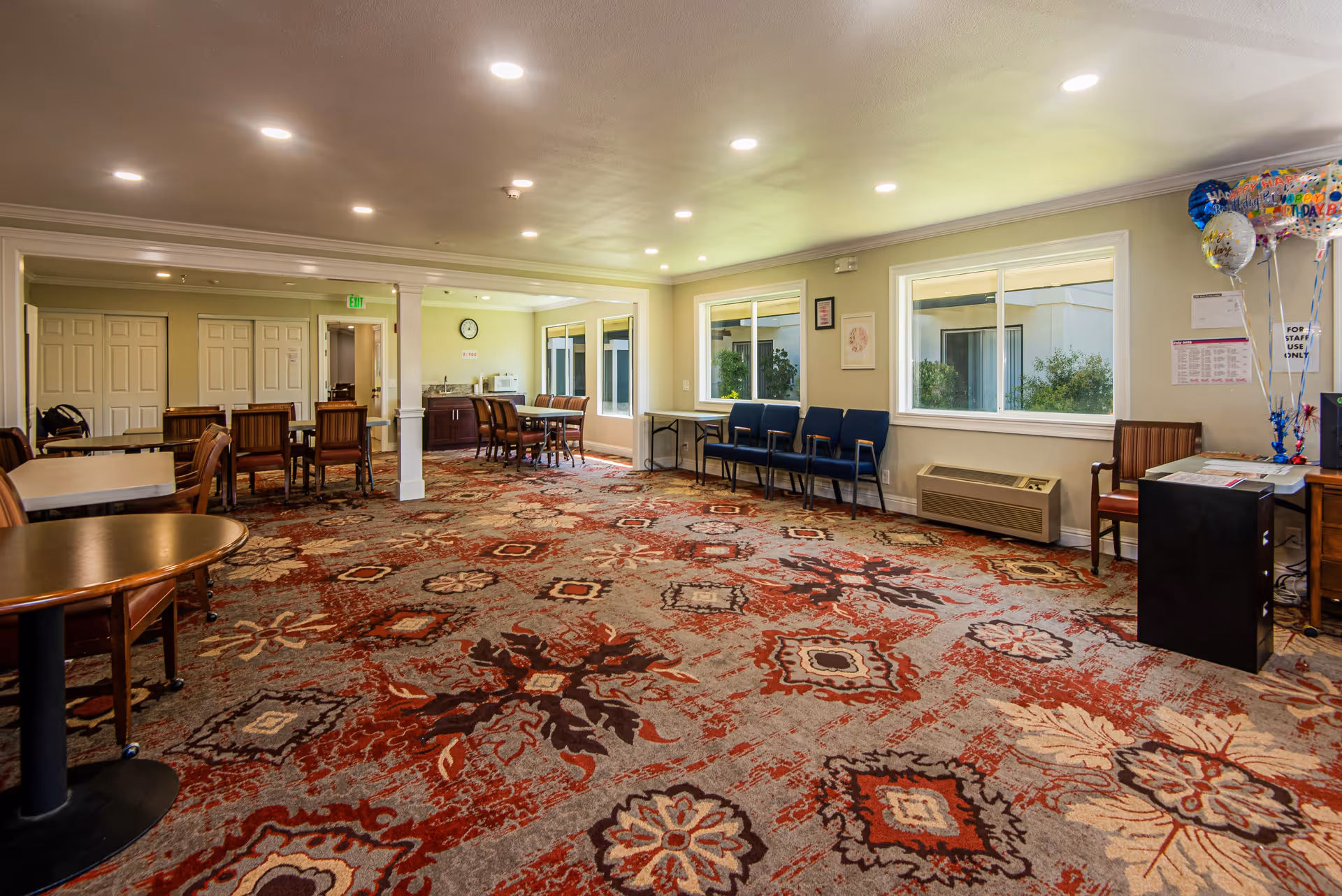 A spacious room with patterned carpet featuring red, beige, and brown colors. The room has several tables and chairs arranged along the walls and in the center. There are large windows letting in natural light, a small kitchenette area with a microwave and sink, and some blue and brown chairs lined up against the walls. Balloons and some papers are visible on a desk in the corner.