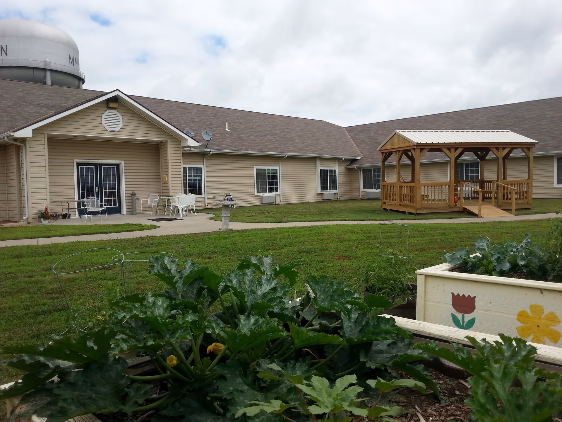 Outdoor view of Marion Assisted Living Community LLC showing a beige building with multiple windows and a covered wooden gazebo on a grassy lawn. In the foreground, there are raised garden beds with green plants and flowers painted on the sides. Patio furniture is visible near the building entrance under a small covered porch.