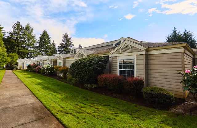 Exterior view of a single-story senior living facility building with beige siding, multiple windows, and a well-maintained lawn with bushes and flowers along the side. A concrete walkway runs parallel to the building, and tall trees are visible in the background under a partly cloudy sky.