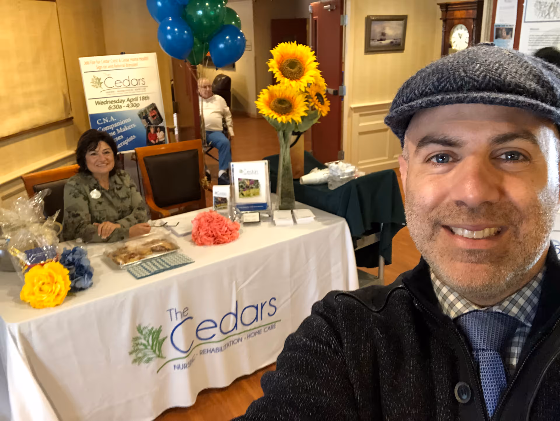 A man wearing a flat cap and tie takes a selfie in front of a table with a white tablecloth that reads 'The Cedars Nursing Rehabilitation Home Care.' Behind the table, a woman sits smiling with balloons and a vase of sunflowers on the table. There is a sign in the background with information about The Cedars.