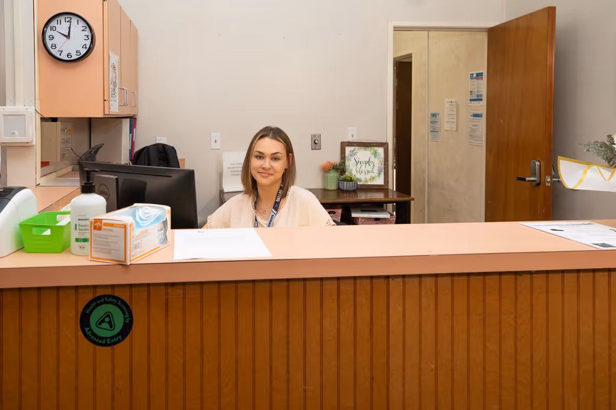 A woman sitting behind a reception desk in an office area with a computer monitor, hand sanitizer, and a box of gloves on the counter. There is a clock on the wall showing 12:05, a door open to a hallway, and a framed sign that says 'Simply Blessed' on a table behind her.