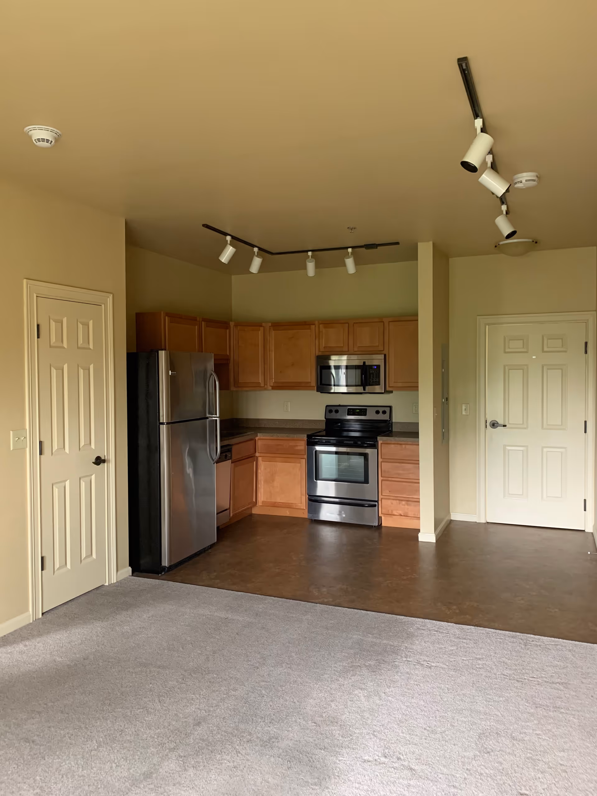 An empty kitchen area in a senior living facility featuring wooden cabinets, a stainless steel refrigerator, stove, microwave, and dishwasher. The kitchen has a brown floor and is adjacent to a carpeted living space. There are two closed doors visible, one on the left and one on the right side of the kitchen. Ceiling track lighting is installed above the kitchen area.