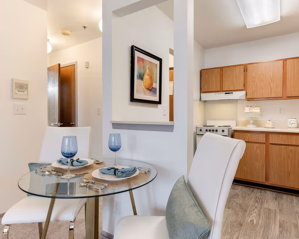 A small dining area with a round glass table set for two with blue wine glasses, plates, and napkins. Two white cushioned chairs with light blue pillows are placed around the table. The dining area is adjacent to a kitchen with wooden cabinets, a stove, and a countertop with decorative items. A framed painting of a pear hangs on the wall between the dining area and kitchen.