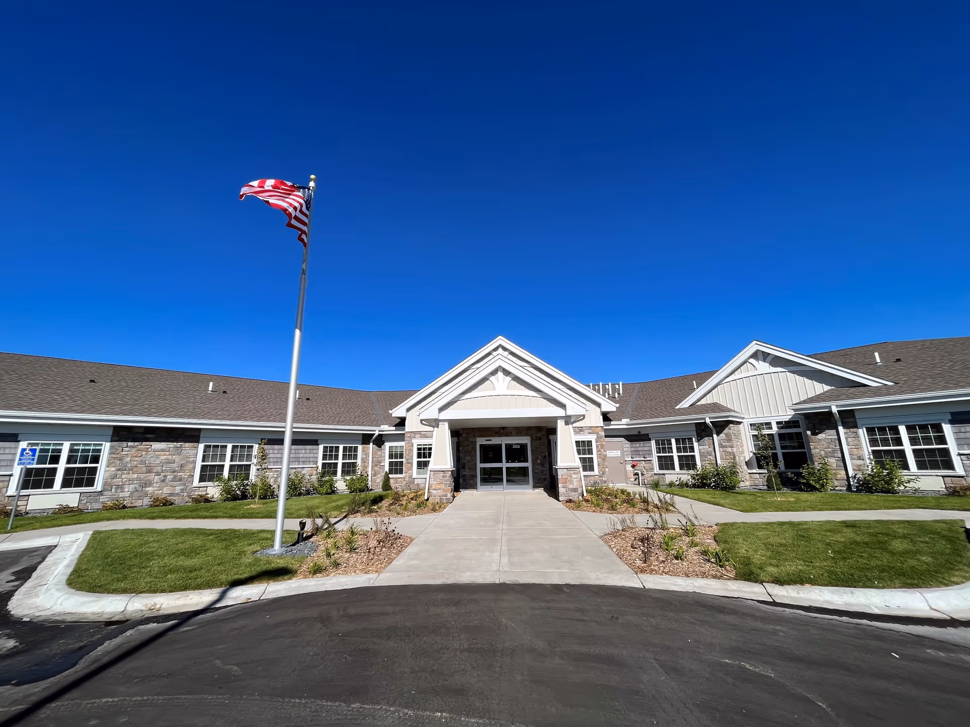Front exterior view of NorBella Senior Living - Centerville building with a clear blue sky, an American flag on a flagpole, a paved driveway, and landscaped green grass and plants.