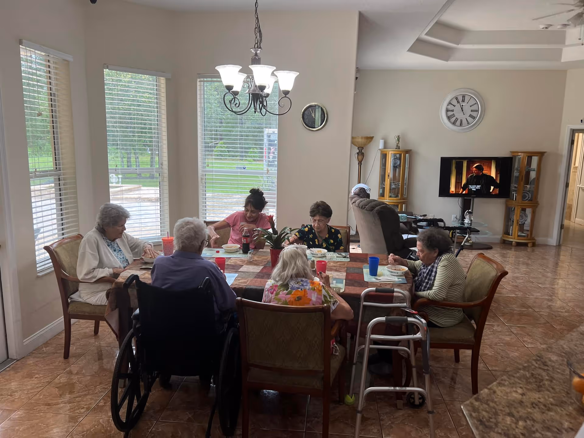 A group of elderly people sitting around a dining table in a well-lit room with large windows. They are eating and socializing. The room has tiled floors, a chandelier, and a television mounted on the wall in the background. Some mobility aids like a wheelchair and a walker are visible.