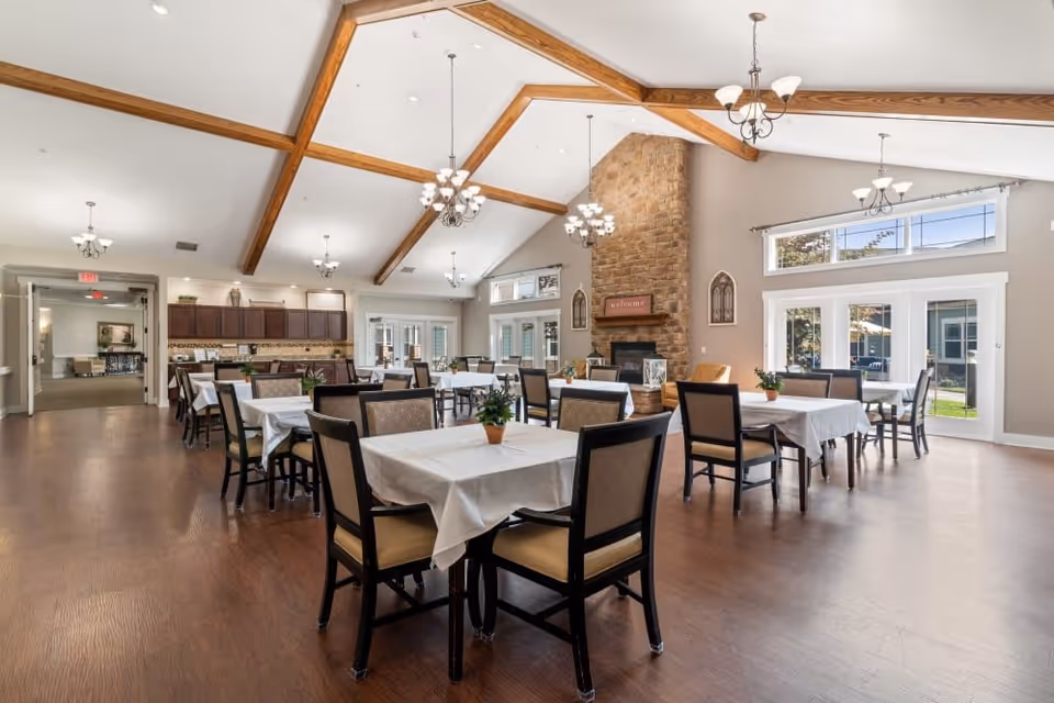 A spacious dining room with multiple tables covered in white tablecloths, each surrounded by chairs. The room features a high vaulted ceiling with wooden beams and several chandeliers. There is a large stone fireplace with a 'welcome' sign above it, and large windows and glass doors letting in natural light. The floor is wooden, and there are small potted plants on each table.