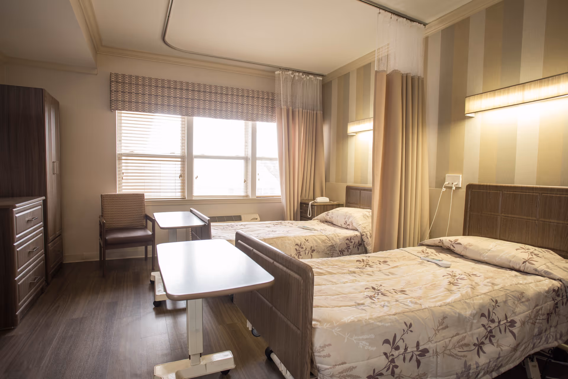 A well-lit room with two single beds separated by a curtain. Each bed has a floral patterned bedspread and a headboard with a wall-mounted light above. There are two overbed tables, a wooden chair, a wooden dresser, and a large window with blinds and a valance letting in natural light.