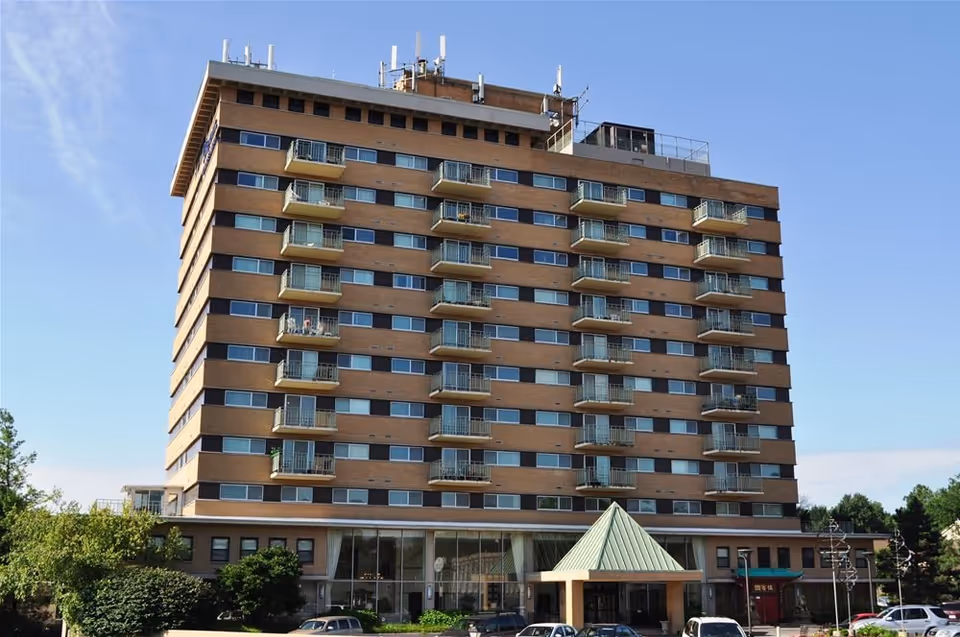 Exterior view of a multi-story brick building with balconies on each floor under a clear blue sky. The building has a green peaked entrance canopy and several cars parked in front. Trees and shrubs surround the building.