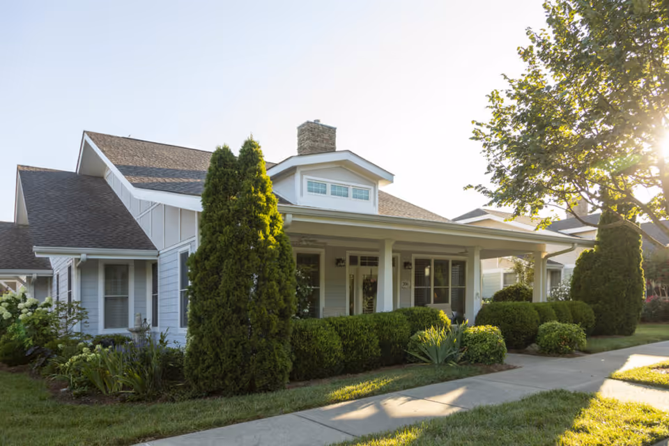 Exterior view of a single-story residential building with a covered front porch, surrounded by well-maintained bushes, trees, and a sidewalk in front. The sun is shining from the right side, casting shadows on the lawn and walkway.