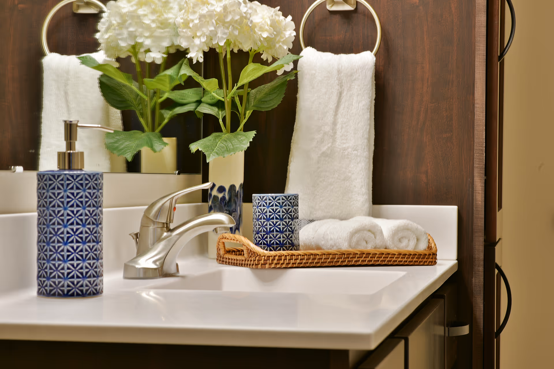 Bathroom vanity with a chrome faucet, patterned soap dispenser, wicker tray holding rolled towels and a small floral arrangement.