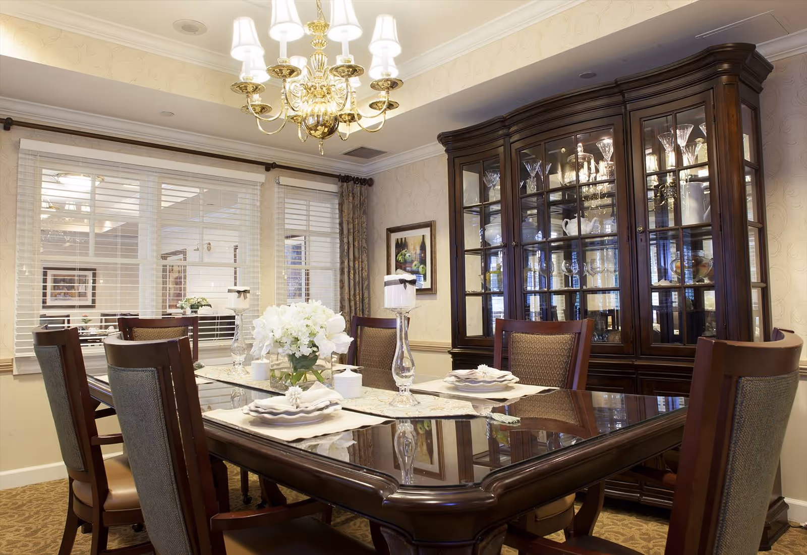 A formal dining room with a dark wooden dining table set for six, featuring white plates, napkins, and a centerpiece of white flowers. Behind the table is a large dark wooden china cabinet displaying glassware and dishes. A gold chandelier hangs from the ceiling, and there are windows with white blinds and patterned curtains on the left wall.
