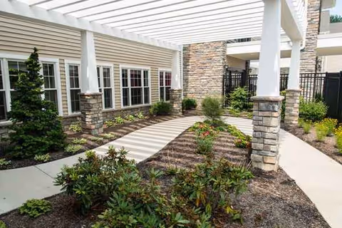Covered outdoor courtyard with stone columns, walkway, and landscaped shrubs beside a beige building.