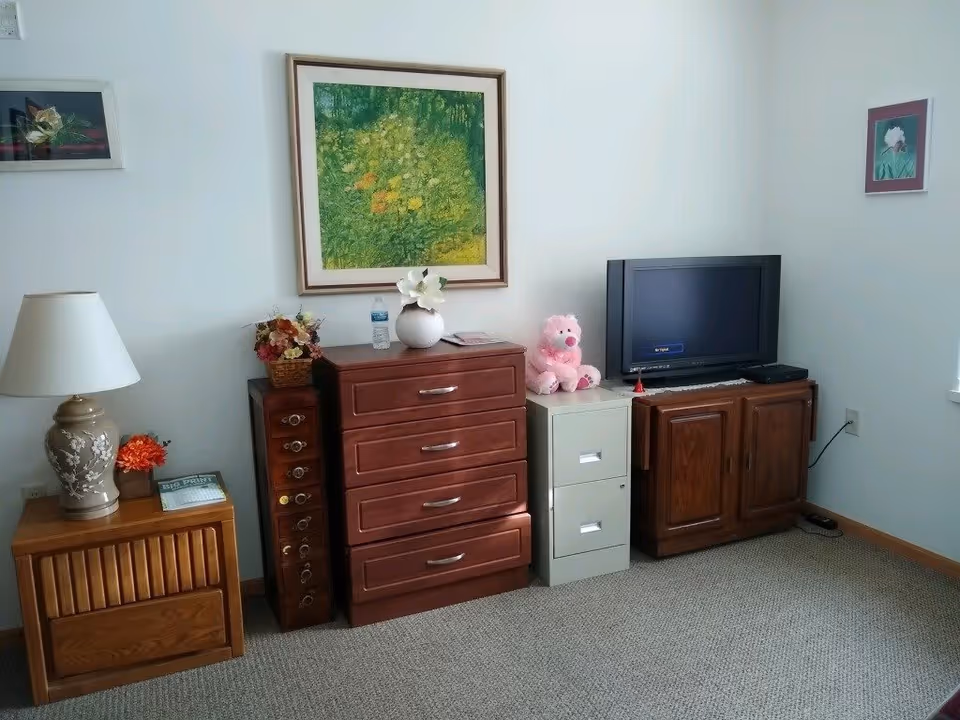 A corner of a senior living room with a wooden nightstand holding a lamp and a small flower arrangement, a wooden chest of drawers with a vase of flowers and a water bottle on top, a small vertical wine rack, a white two-drawer filing cabinet with a pink teddy bear on top, and a wooden TV stand with a flat-screen TV. The walls are white with three framed pictures hanging.