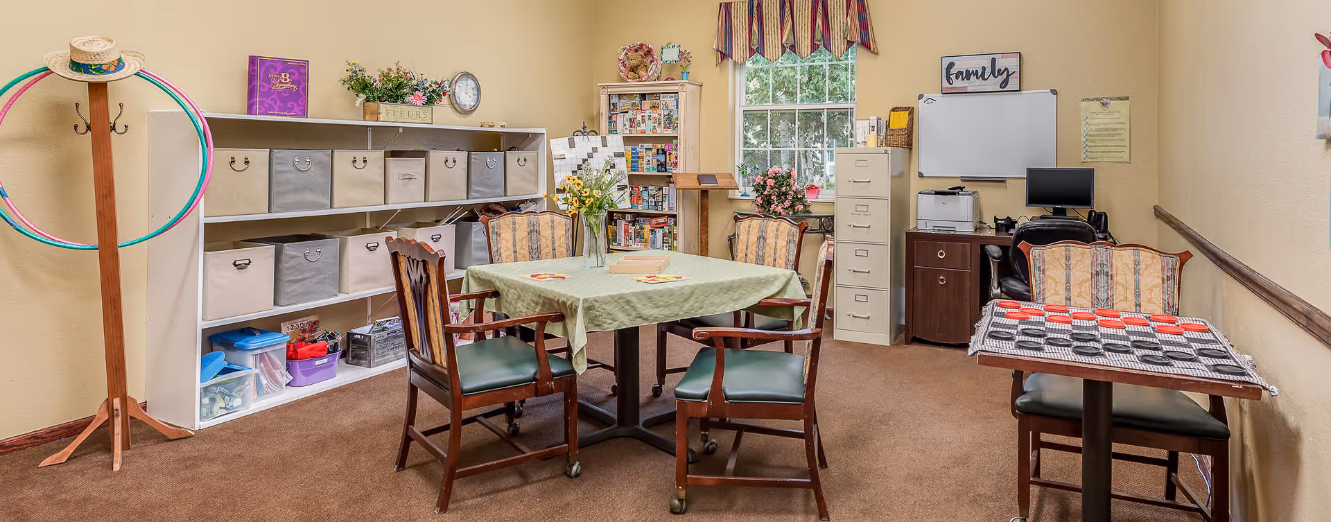 A cozy activity room with a table covered by a green tablecloth surrounded by four chairs. Another smaller table with a checkerboard game is to the right. Shelves with storage bins and various items are against the left wall. A coat rack with hula hoops and a hat stands in the corner. A window with striped valance lets in natural light. A filing cabinet, computer, printer, and whiteboard are against the far wall. The room has beige walls and brown carpet.