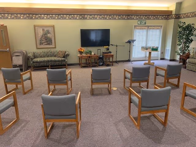 A community room with several wooden chairs arranged in rows facing a wall-mounted flat screen TV. There is a floral-patterned couch along the left wall, a small table with flowers and decorations beneath the TV, and a music stand near a door with a 'No Exit' sign. The room has carpeted flooring and light green walls with a floral border near the ceiling.