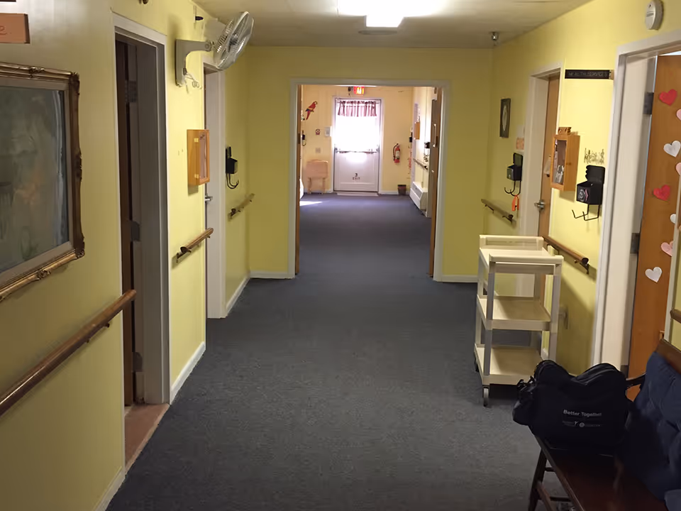 A long hallway in a senior living facility with yellow walls and gray carpet. There are handrails on both sides of the hallway, a wall-mounted fan, a painting on the left wall, and a small white rolling cart on the right. At the end of the hallway, there is a door with a window letting in natural light. A bench with a black bag on it is visible on the right side near the foreground.
