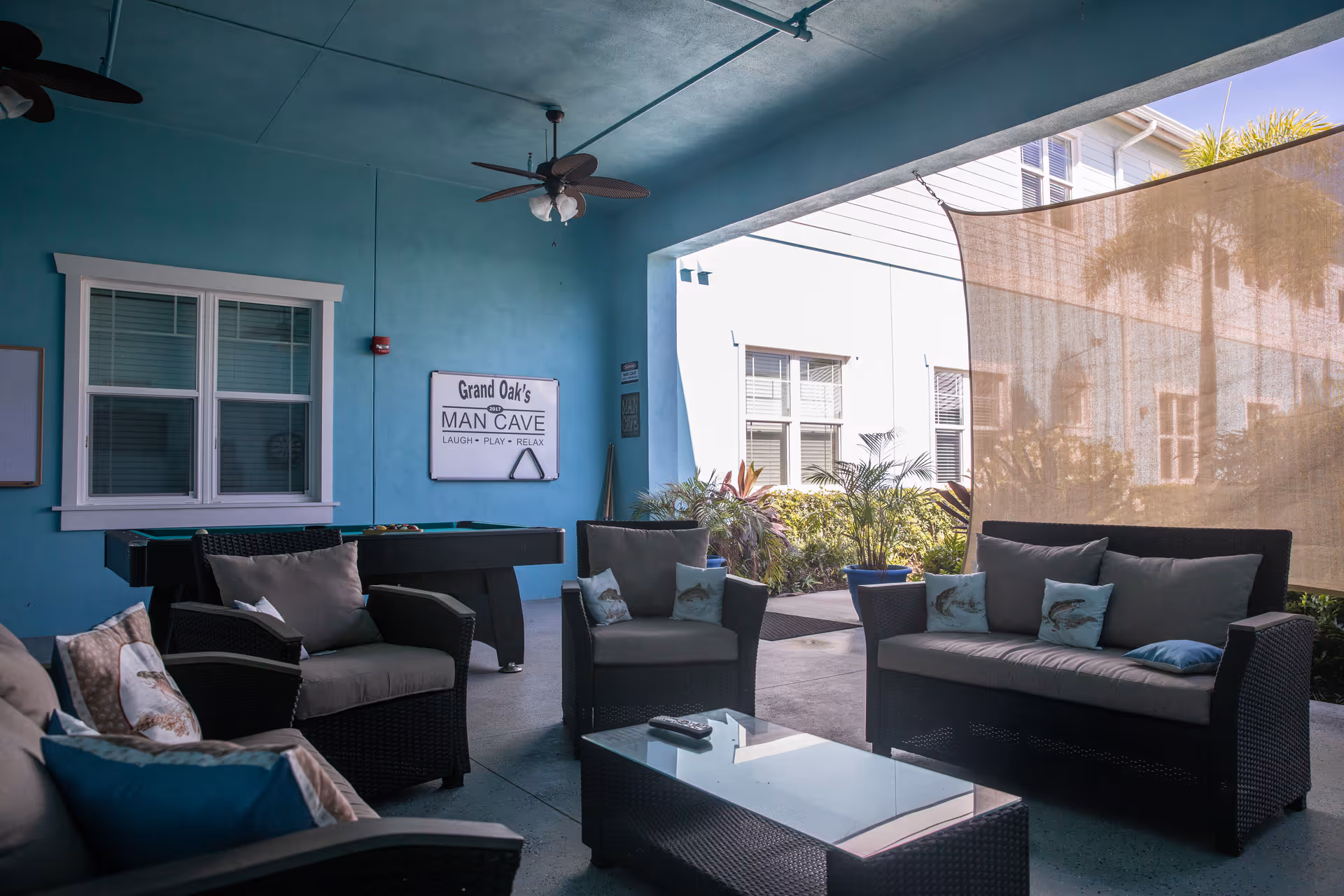 Covered outdoor seating area with wicker furniture including a sofa, armchairs, and a glass-top coffee table. There is a pool table against a blue wall with a sign that reads 'Grand Oak's MAN CAVE LAUGH - PLAY - RELAX'. Ceiling fans are mounted on the ceiling, and plants are visible outside the covered area.