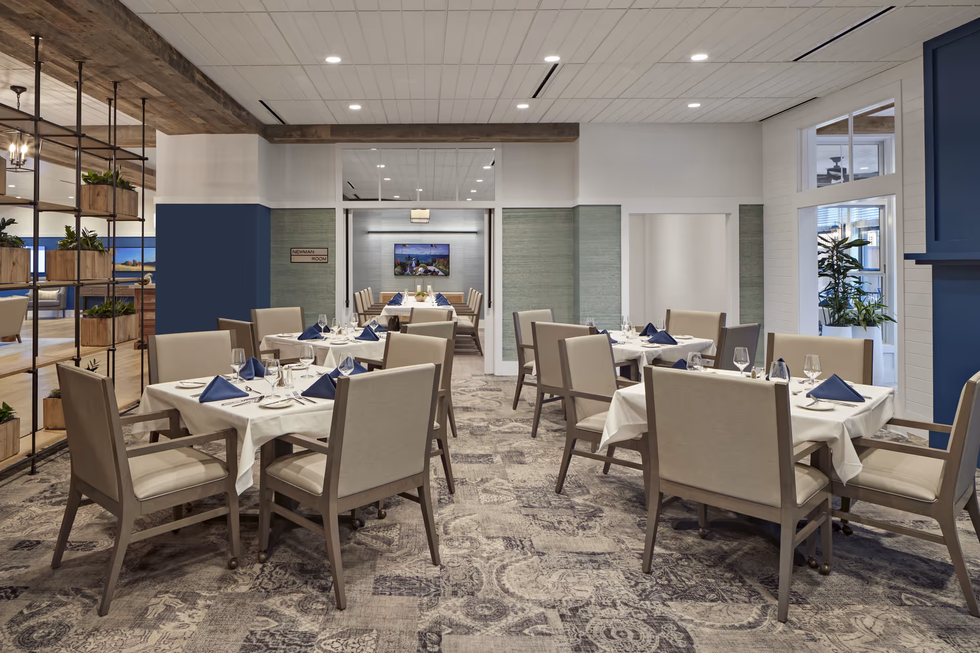 A well-lit dining room with several tables covered in white tablecloths, each set with blue napkins, glassware, and silverware. The room features beige cushioned chairs, a patterned carpet, and a decorative partition with plants on the left side. In the background, there is a smaller room labeled 'Newman Room' with a long table and chairs, and a TV mounted on the wall.