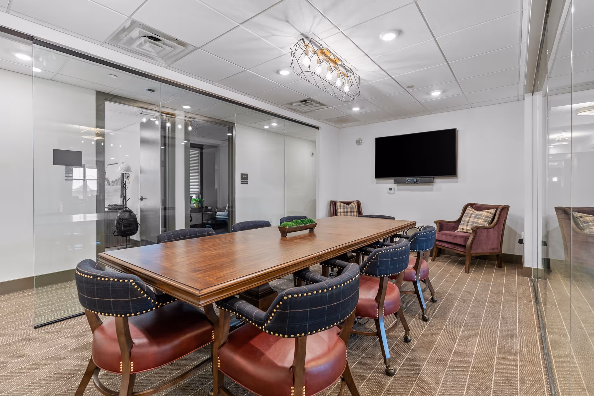 A modern conference room with a long wooden table surrounded by eight upholstered chairs with plaid backs and red seats. The room has glass walls, a flat-screen TV mounted on the wall, two armchairs with plaid cushions in the corner, and a decorative light fixture on the ceiling.