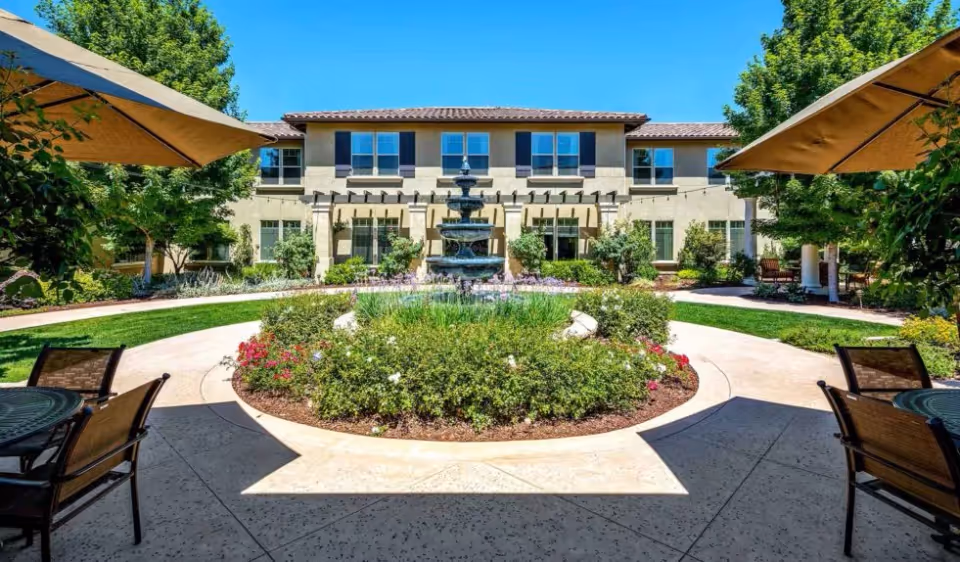 Sunny courtyard with a central multi-tiered fountain surrounded by a circular flowerbed, patio tables with umbrellas, and a two-story building in the background.