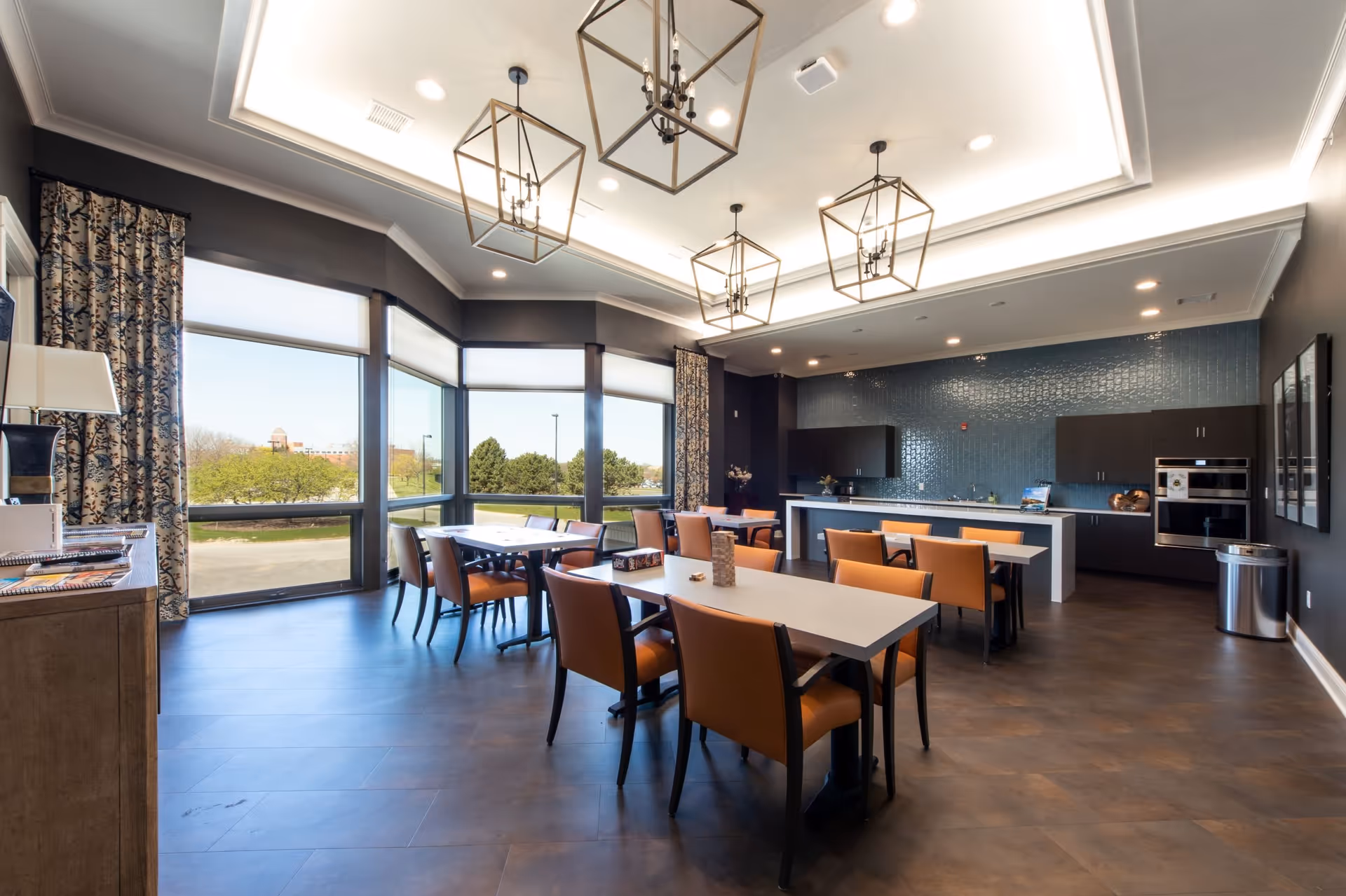 A spacious dining room with several tables and orange cushioned chairs arranged neatly. Large windows with patterned curtains allow natural light to fill the room, showing a view of trees and a building outside. The ceiling features modern geometric pendant lights, and the back wall has a blue tiled backsplash with dark cabinetry and built-in ovens. The floor is covered with dark wood-like tiles.