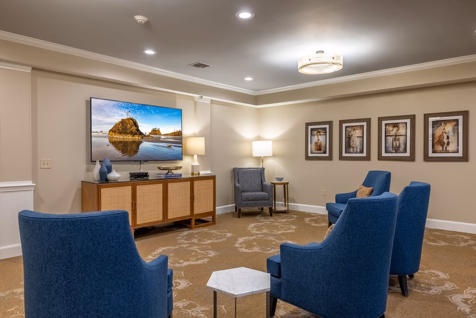 A cozy living room area with four blue upholstered chairs arranged around a small white hexagonal table. A wooden cabinet with a large flat-screen TV mounted above it is against the wall. The TV displays a scenic beach with rock formations. There are two table lamps providing warm lighting, and five framed pictures hang on the beige wall. The room has a patterned carpet and recessed ceiling lights.