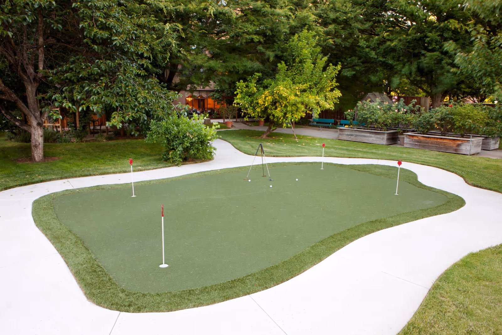 A small outdoor putting green with four holes marked by red flags, surrounded by a concrete pathway. The area is lush with green grass, trees, and raised garden beds in the background, suggesting a peaceful garden setting.