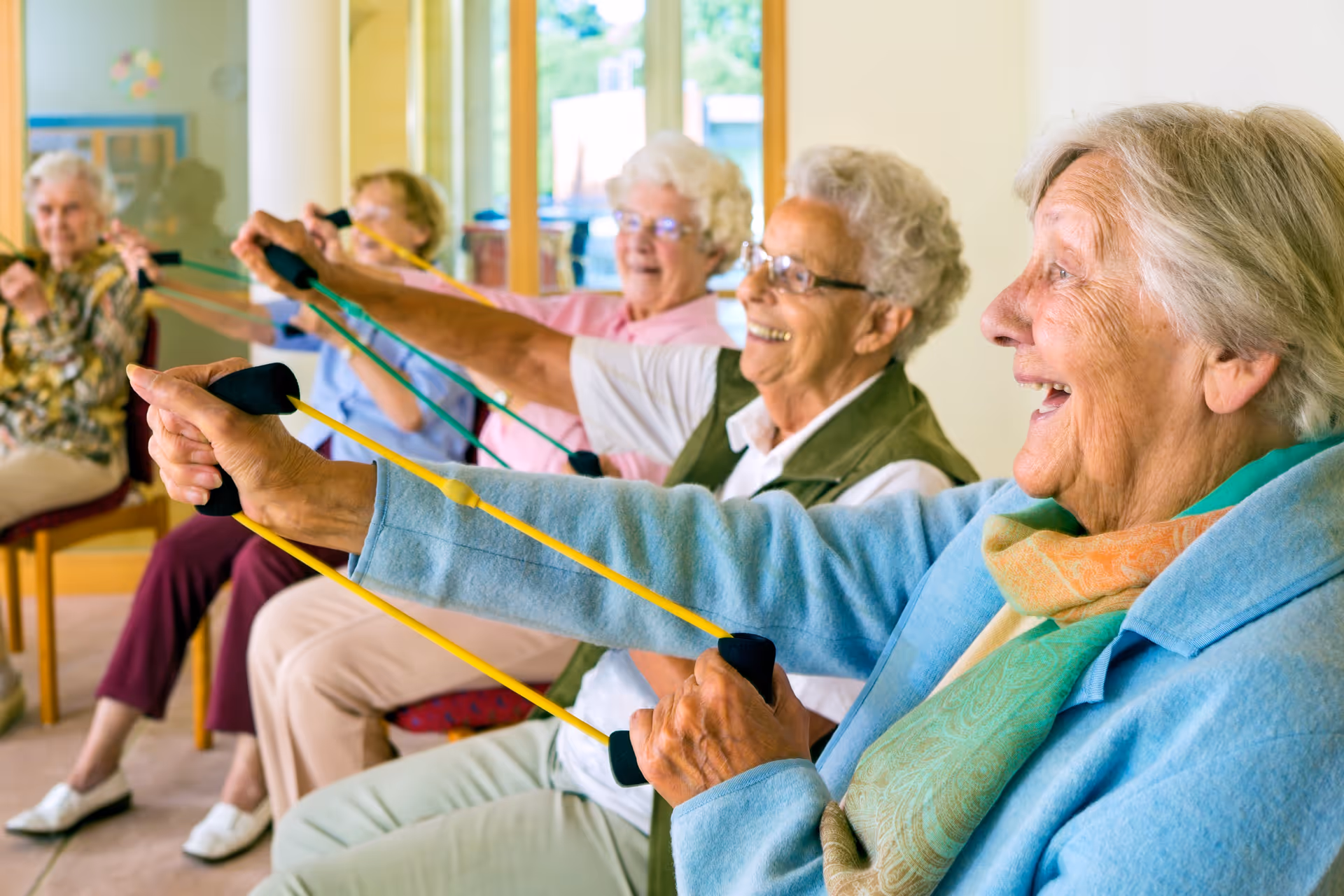 A group of elderly women sitting in chairs indoors, smiling and exercising with resistance bands stretched forward.