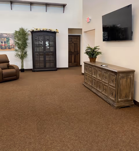 Spacious senior living lounge with a wall-mounted TV, wooden sideboard and glass display cabinet, a recliner, and potted plants.
