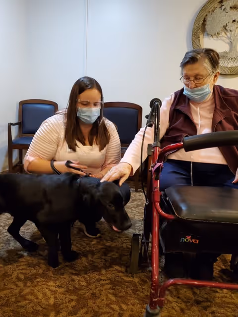 An elderly woman wearing a mask and sitting with a red walker is petting a black dog. A younger woman, also wearing a mask and glasses, is crouched beside the dog, holding its leash. They are indoors in a room with carpeted floor and chairs against the wall.